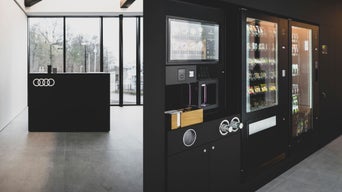 Selbstbedienungsbereich mit Snack- und Getränkeautomaten, daneben schwarzer Tresen mit Audi-Logo vor Glasfront.