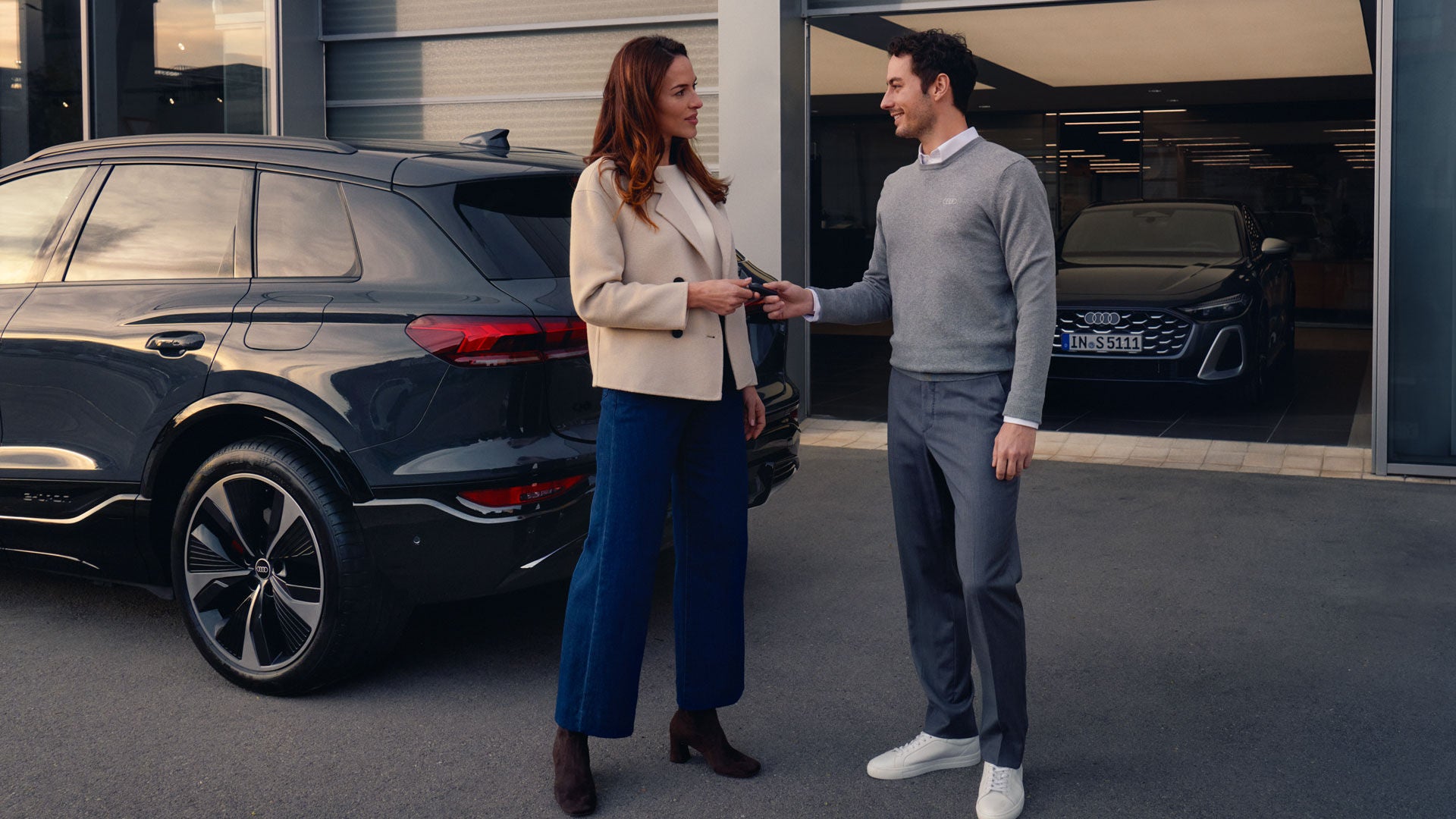 Frau und Mann stehen vor einem Autohaus neben einem schwarzen Audi und übergeben einen Autoschlüssel.