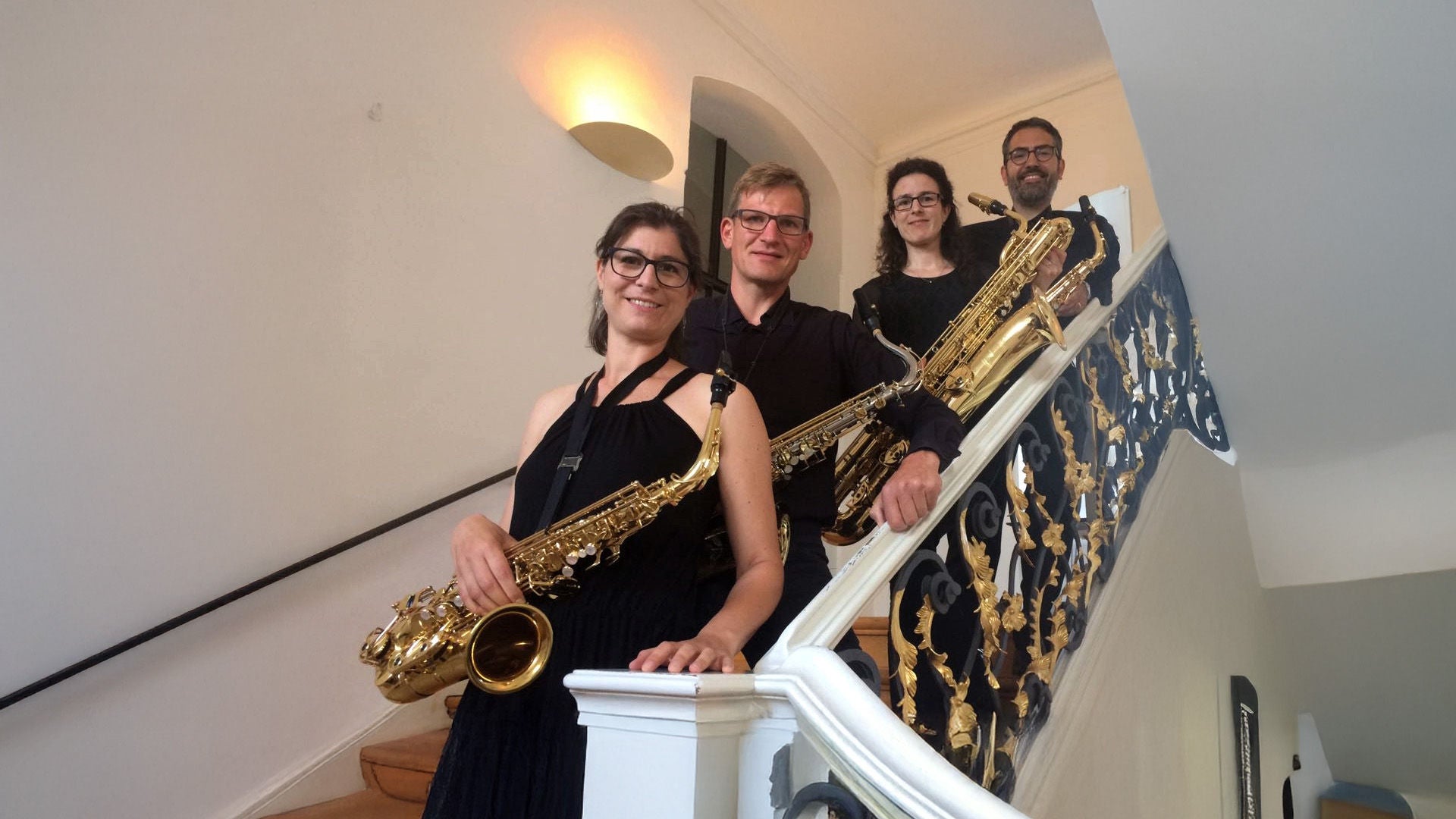 Vier Musikerinnen und Musiker mit Saxophonen stehen lächelnd auf einer Treppe in einem hellen Innenraum.
