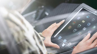 Person bedient ein Tablet mit mehreren App-Symbolen im Innenraum eines Fahrzeugs bei einfallendem Sonnenlicht.