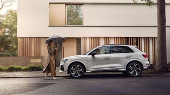 Frau mit Regenschirm geht an geparktem weißen Audi Q3 vor einem modernen Wohnhaus entlang.