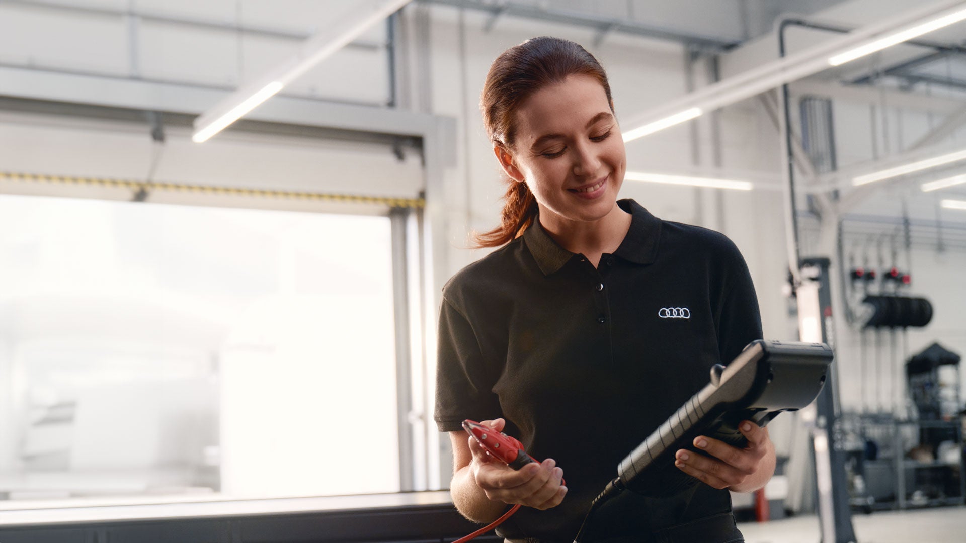 Mitarbeiterin im Audi Poloshirt hält in einer Werkstatt ein Prüfgerät und Batterieklemmen in der Hand.