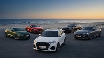 Fünf Audi-Modelle in Rot, Schwarz und Weiß und dunkelblau stehen versetzt hinter und voreinander vor einer Meerkulisse, darunter Schriftzug „Audi Gebrauchtwagen :plus Sommerdeals“.
