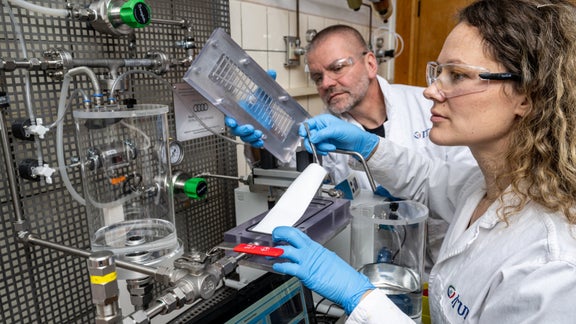 Ein Mann und eine Frau in weissen Laborkitteln und Schutzbrillen betrachten in einem Labor eine Metallplatte.