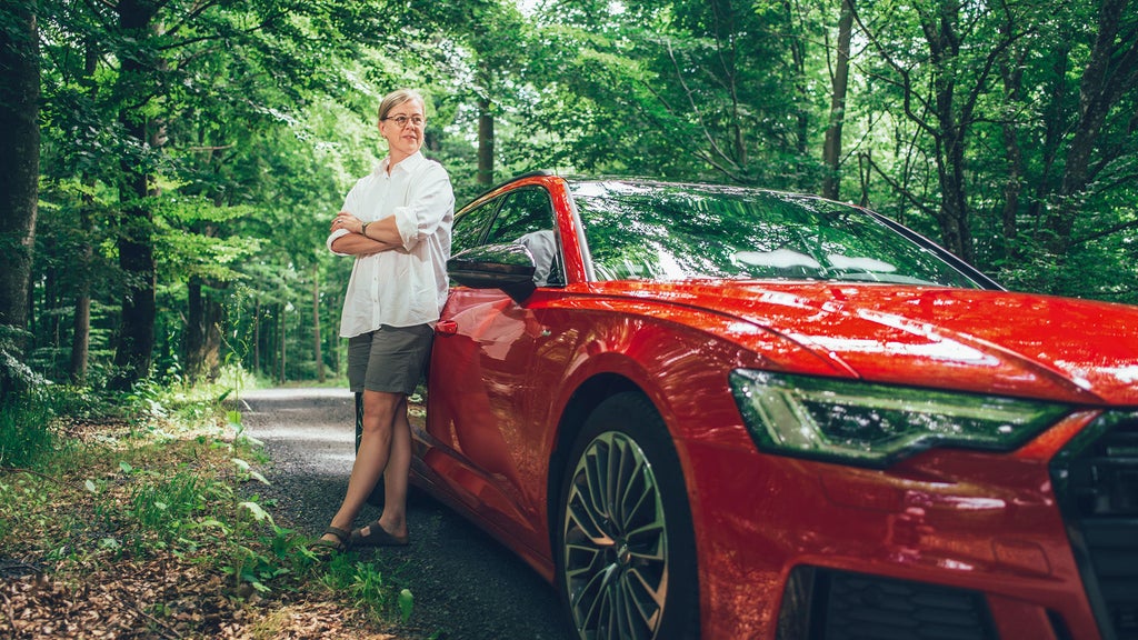 Ulrika Hasselgren lutar sig mot sin röda Audi A6. Bilen står parkerad längs en grusväg i en ljus lövskog