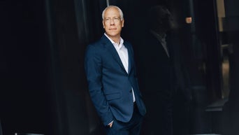 Peter Gladbach steht im blauen Anzug vor dunklem Hintergrund und blickt ruhig in die Ferne.