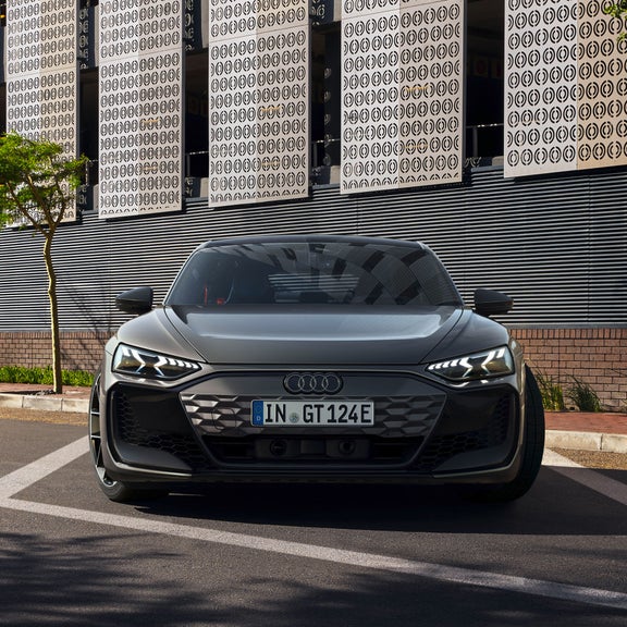 Ein grauer Audi RS e-tron GT steht frontal auf einer Straße mit moderner Metallfassade und Bäumen im Hintergrund.