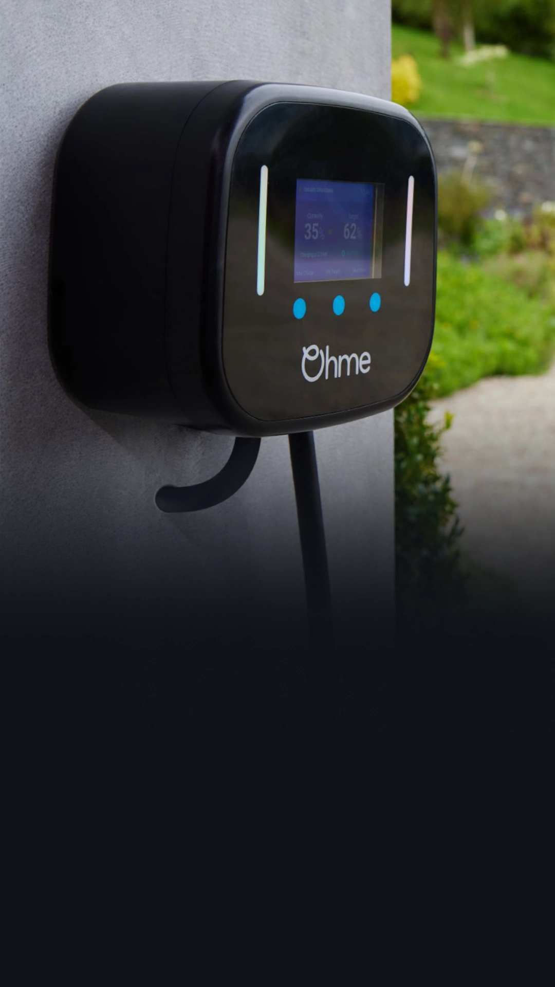 Black Ohme charger on wall with screen displaying 35% and 62%, blue buttons, and white logo; background shows blurred garden and stone wall.