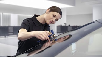 Frau im schwarzen Polo-Shirt repariert konzentriert eine Autoscheibe in heller, moderner Werkstatt.