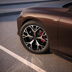 Close-up of the stylish wheel of a brown Audi, featuring a sleek design and red brake callipers, parked on a textured surface.