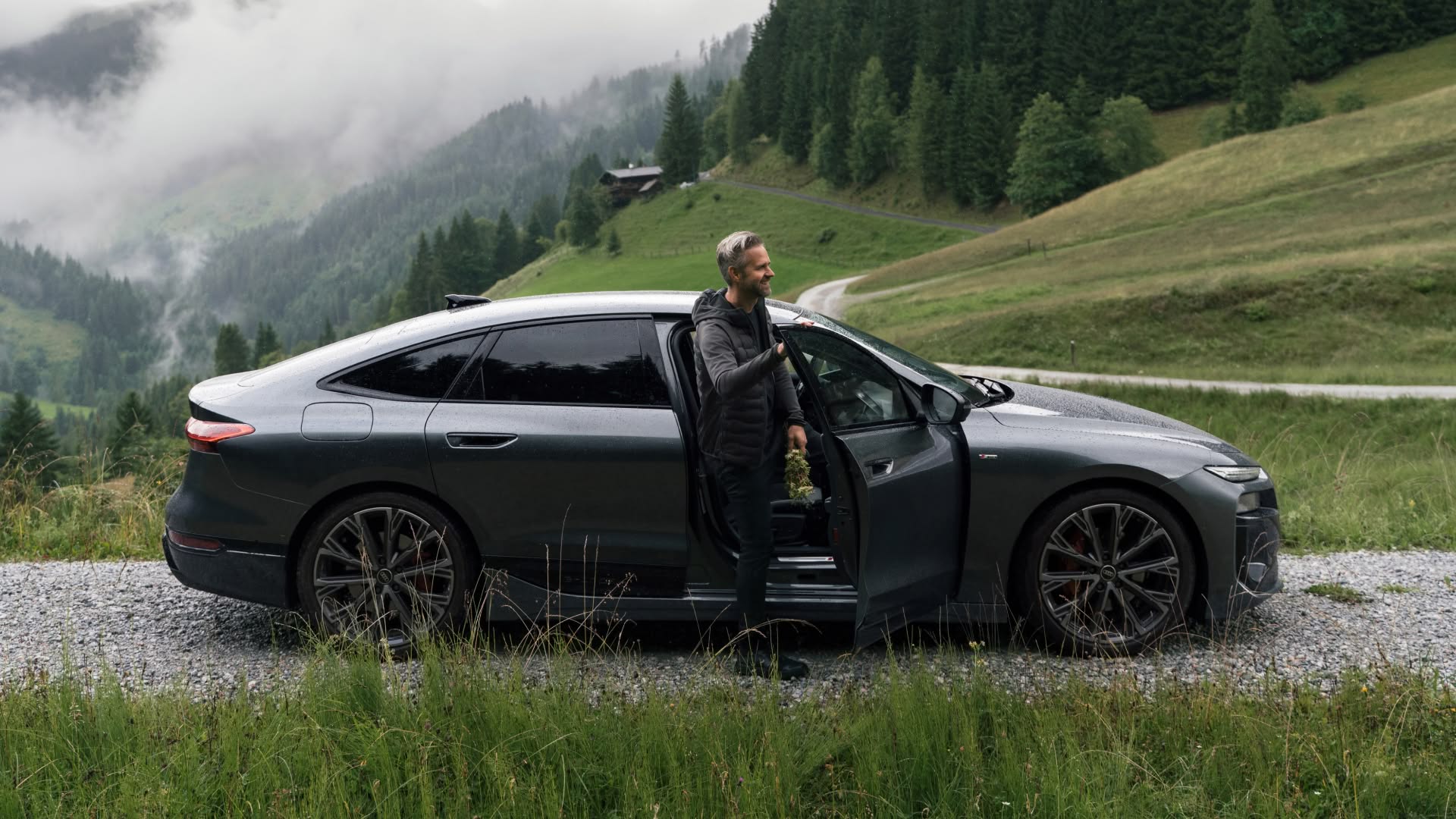 Winkler stoi w otwartych drzwiach Audi A6 Sportback e-tron. Samochód jest zaparkowany na szutrowej drodze, w tle widać łąki, góry i lasy.