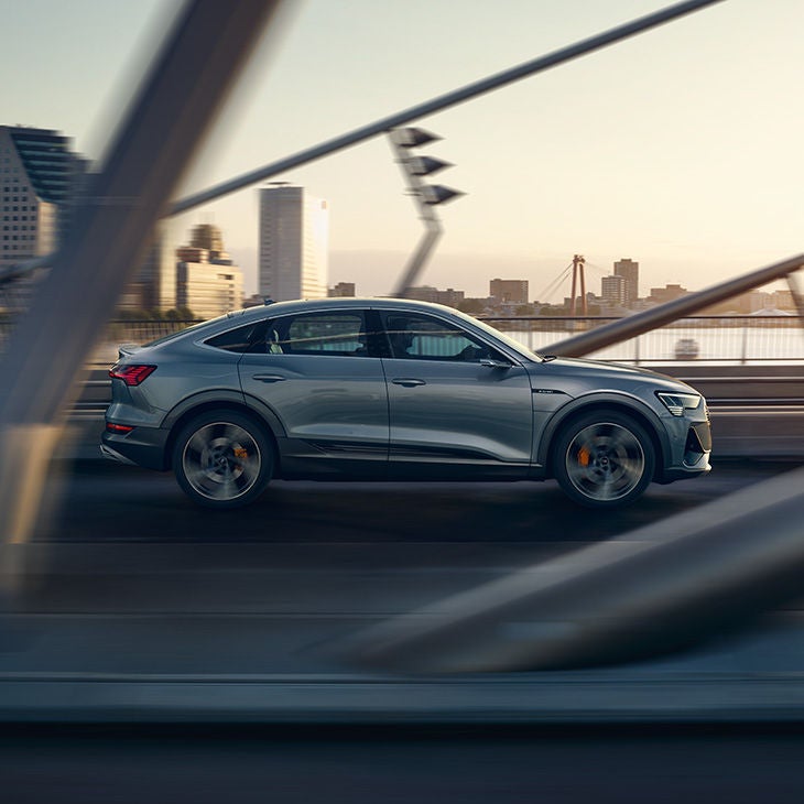 Silberner Audi e-tron Sportback fährt über eine Brücke vor städtischer Skyline bei Sonnenuntergang, seitlich aufgenommen.