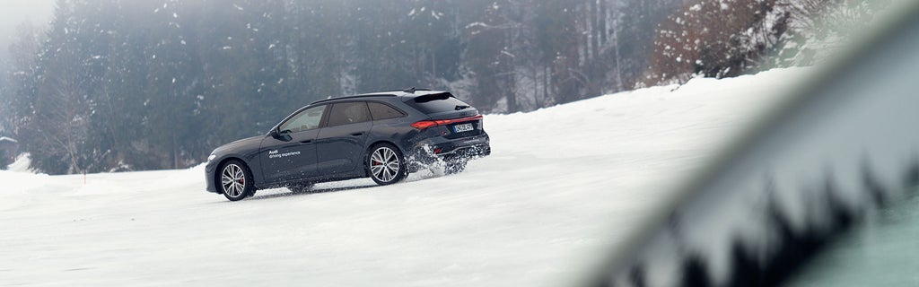 Dunkler Audi S5 Avant fährt auf verschneiter Strecke, wirbelt Schnee auf, dichter Wald und Nebel im Hintergrund.
