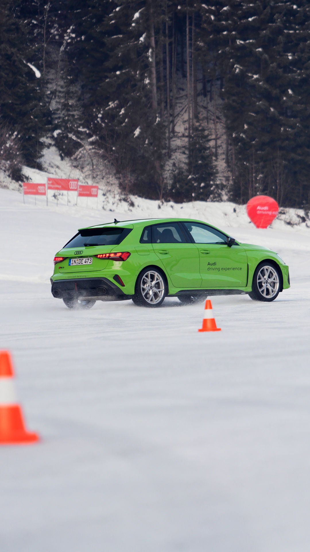 Light green Audi RS 3 Sportback driving on a snow-covered track between orange cones, snow-covered forest in the background.