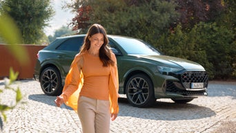 Une femme vêtue d'orange traverse un parvis pavé. À l'arrière-plan, un SUV Audi vert olive est garé devant des arbres et des buissons, dans un cadre en plein air ensoleillé.