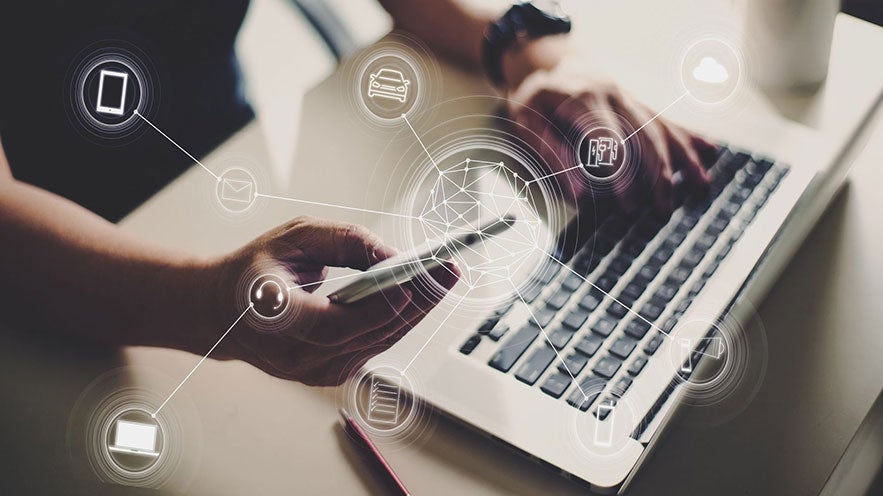 Ein Laptop auf einem Tisch, jemand hat ein Handy in der Hand, darüber ein virtuelles Netz aus Technologie.