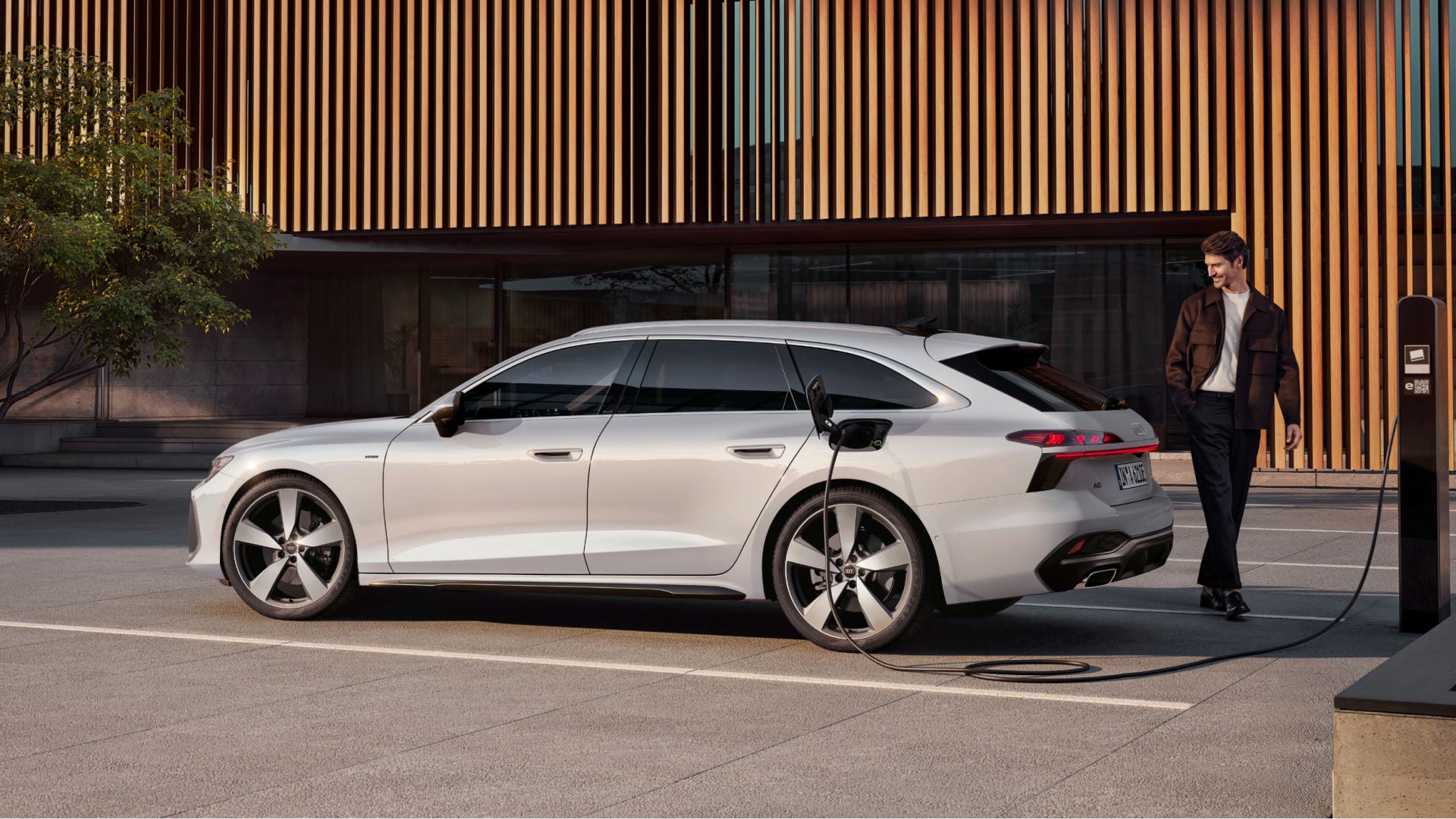 Mann lehnt an einem ladenden, silbernen Audi e-tron Avant vor modernem Gebäude mit Holzfassade.