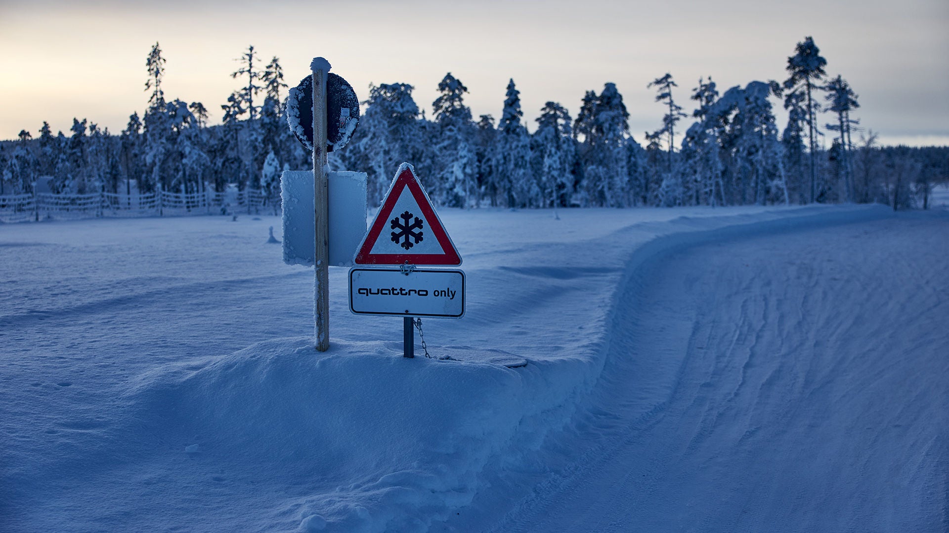 A snow-covered icy road with a warning sign (snowflake) and the note “quattro only,” with a forest in the background.