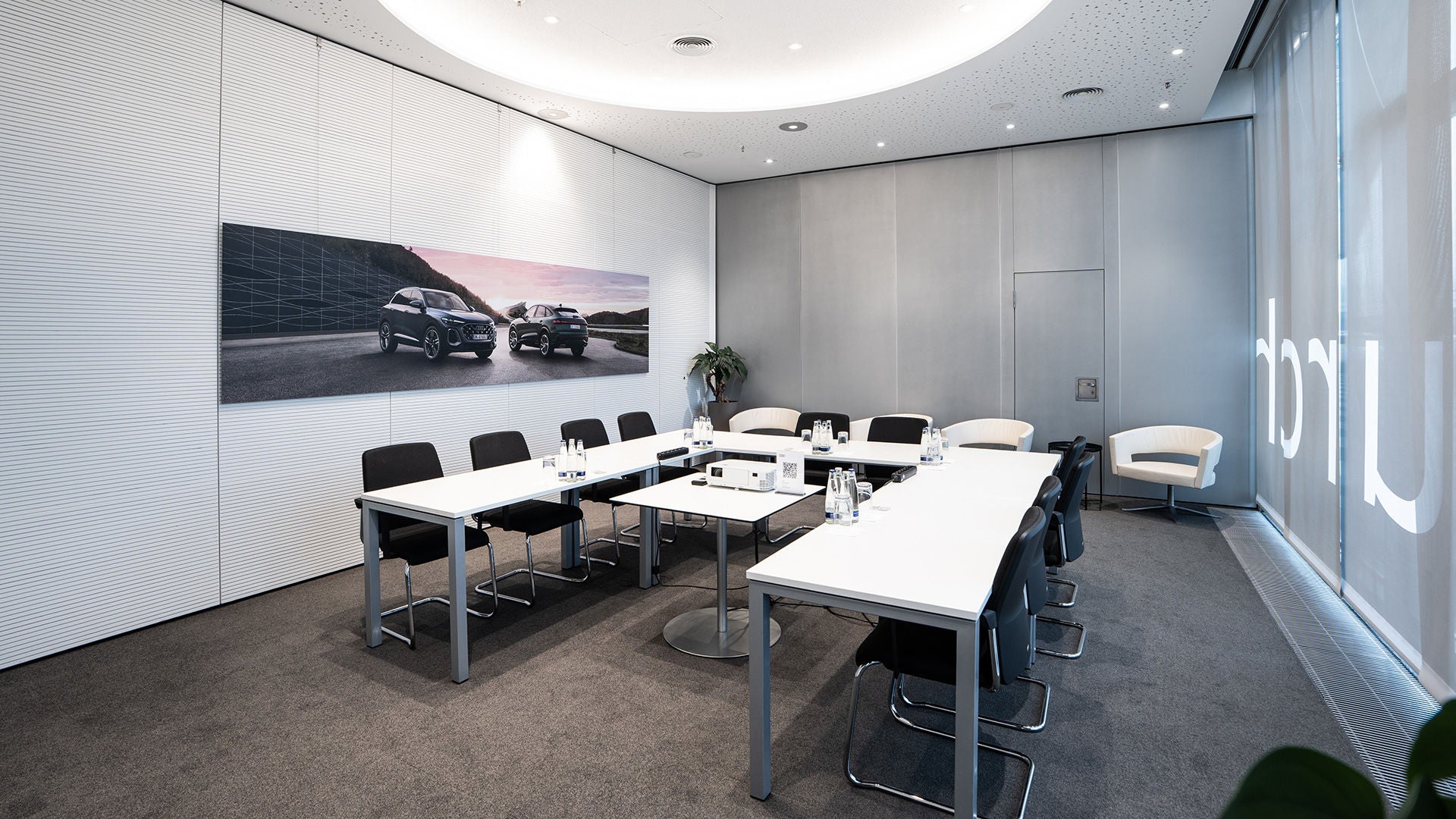 Konferenzraum mit U-förmiger Tischanordnung, Stühlen, Getränken, Projektor und Autowerbung an der Wand.