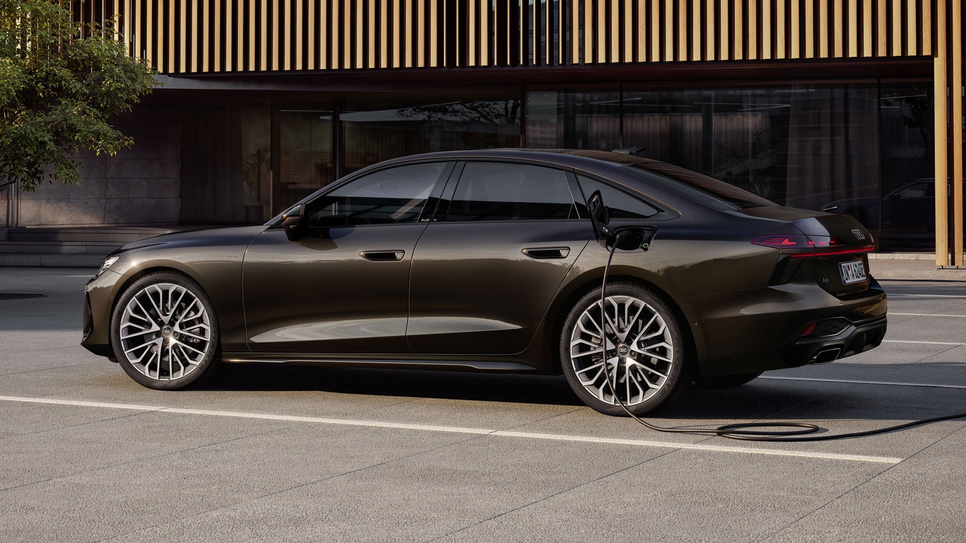 Die A6 Limousine e-hybrid in Havannaschwarz Metallic steht seitlich vor einer Gebäudefassade und ist dabei an eine Ladestation angeschlossen.