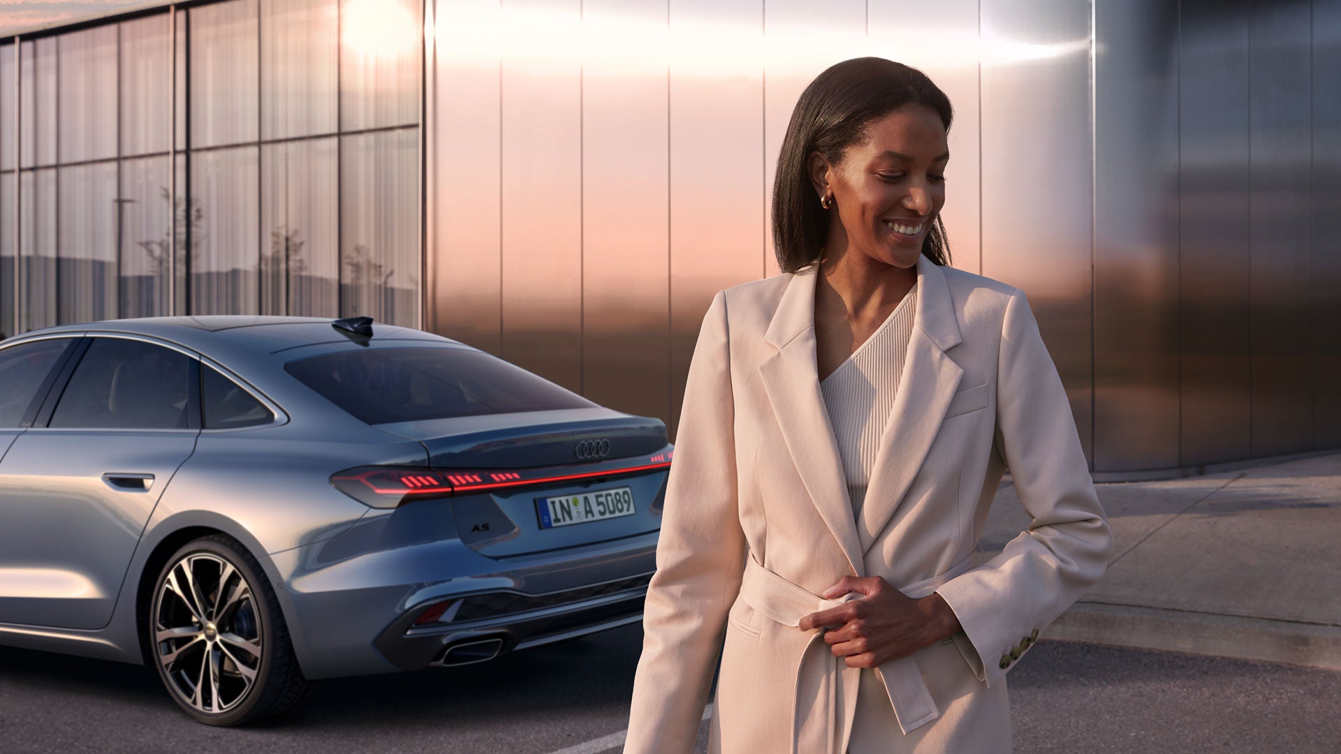 A person in a light suit stands smiling next to a silver Audi A5 car, with a glass building behind them.