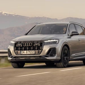 A silver Audi SUV driving on a scenic mountain road under a clear sky, with snow-capped peaks in the distance. The mood is adventurous and serene.