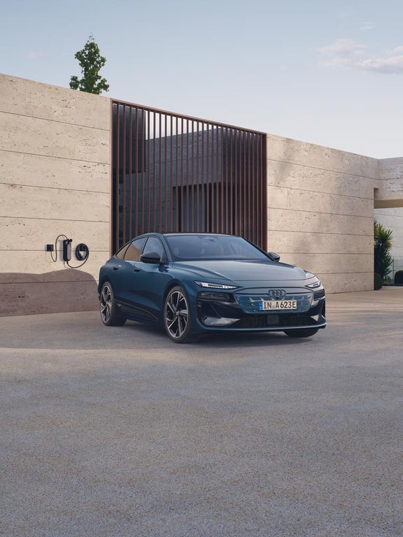 Blauer Audi A6 Sportback e-tron von vorn links, angeschlossen an eine Wandladestation, vor heller Fassade geparkt.