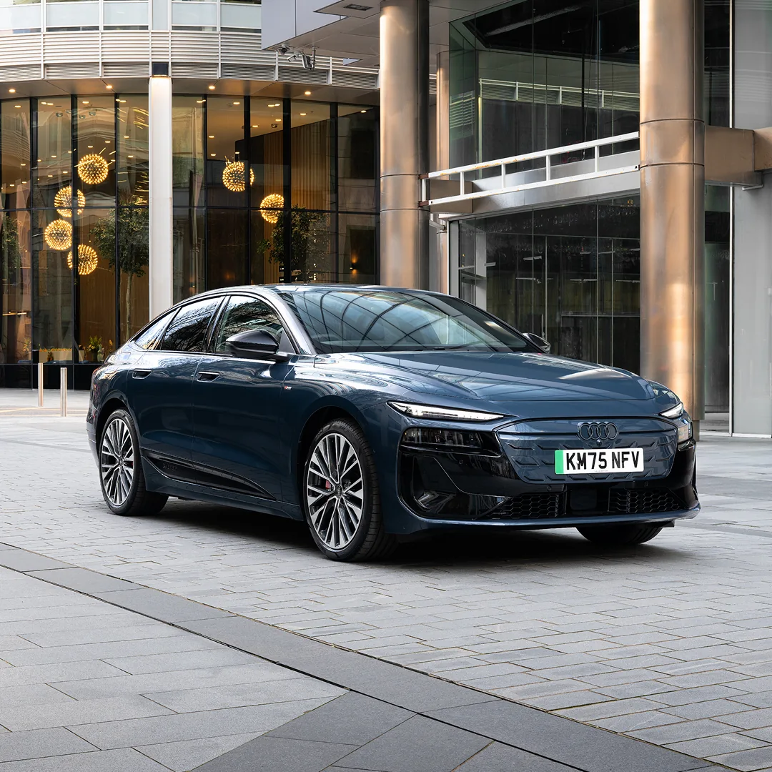 Dark blue Audi e-tron Sportback parked outside a modern office building, shown from the front three-quarter angle. The vehicle features a streamlined fastback design, slim LED headlights, a closed front grille and large multi-spoke alloy wheels. Floor-to-ceiling glass windows, metal columns and decorative pendant lighting create a premium urban backdrop under daylight conditions.