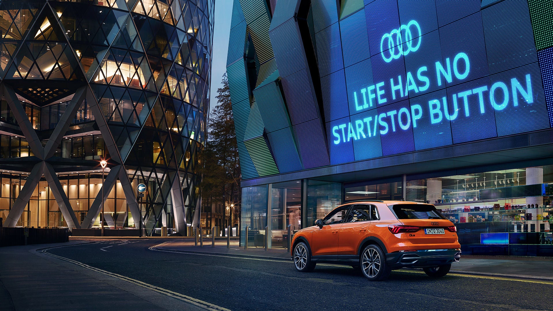 Ein orangefarbener Audi SUV steht nachts vor einem Gebäude mit Leuchtschrift „Life has no start/stop button“ in urbaner Umgebung.