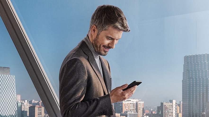 Mann schaut auf sein Handy vor einem großen Fenster, durch das man die Skyline sieht