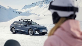 Frau mit Skihelm blickt auf blauen Audi S6 Avant e-tron im Schnee, im Hintergrund verschneite Berglandschaft.