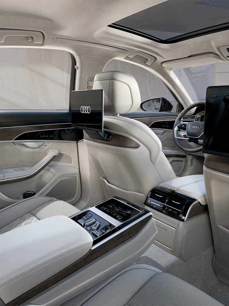 Seitliche Sicht auf die Rückbank, luxuriöser Fond des Audi A8 mit zwei Bildschirmen und heller Lederausstattung. Auf den Bildschirmen ist das Audi Logo.