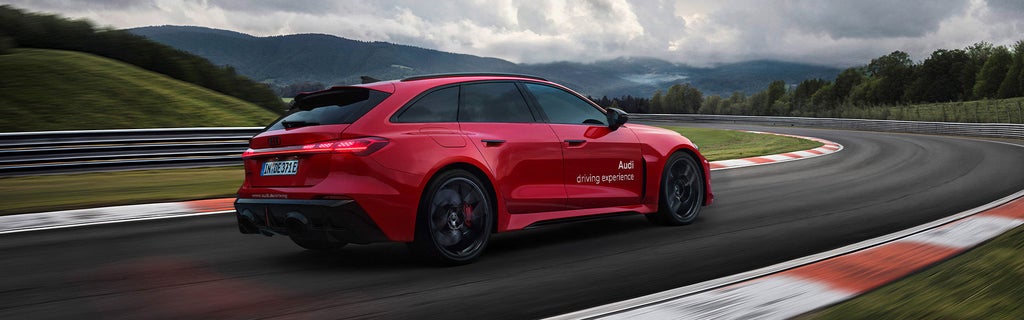 Roter Audi RS 5 Avant fährt auf Rennstrecke durch Kurve, bewölkter Himmel und grüne Hügel im Hintergrund.