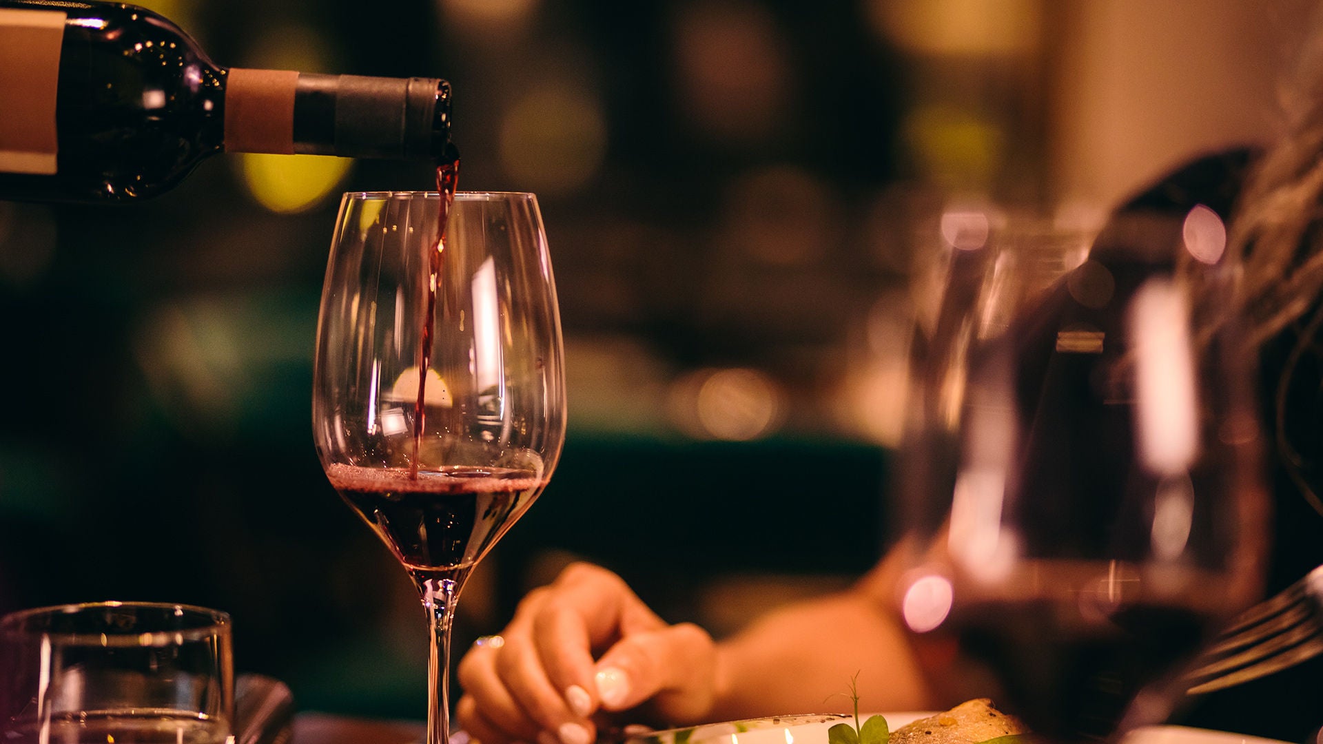 Rotwein wird in ein Weinglas eingeschenkt; im Hintergrund sitzt eine Person an einem warm beleuchteten Esstisch.