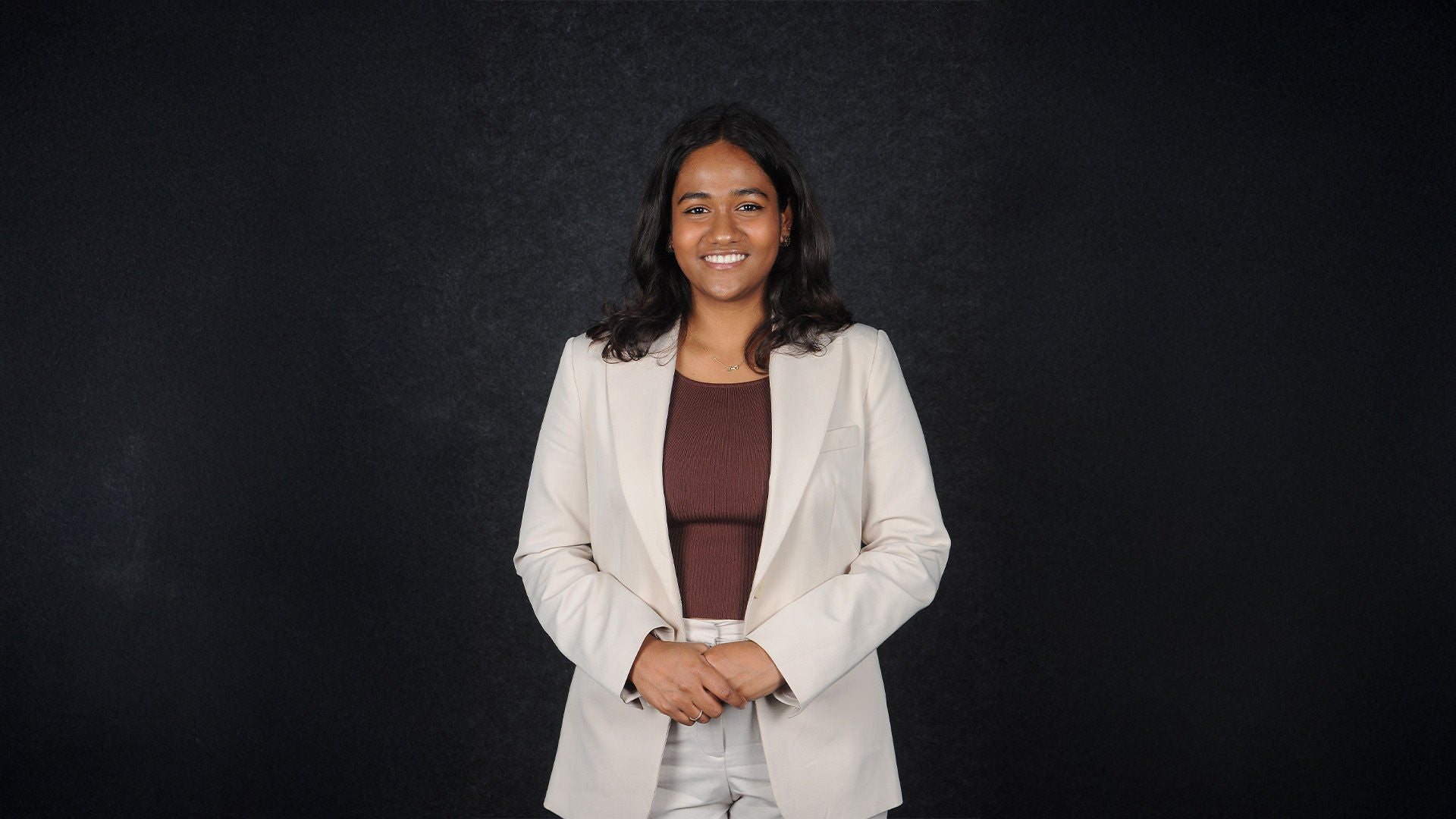 Frau mit mittellangen dunklen Haaren im hellen Blazer, lächelnd, vor schwarzem Hintergrund.