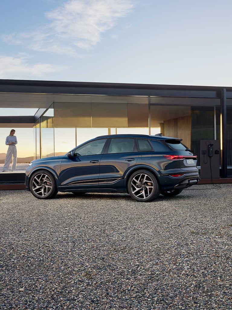 Ein blauer Audi Q6 SUV e-tron vor modernem Haus mit Glasfassade, Frau steht auf Terrasse, bergige Landschaft im Hintergrund.