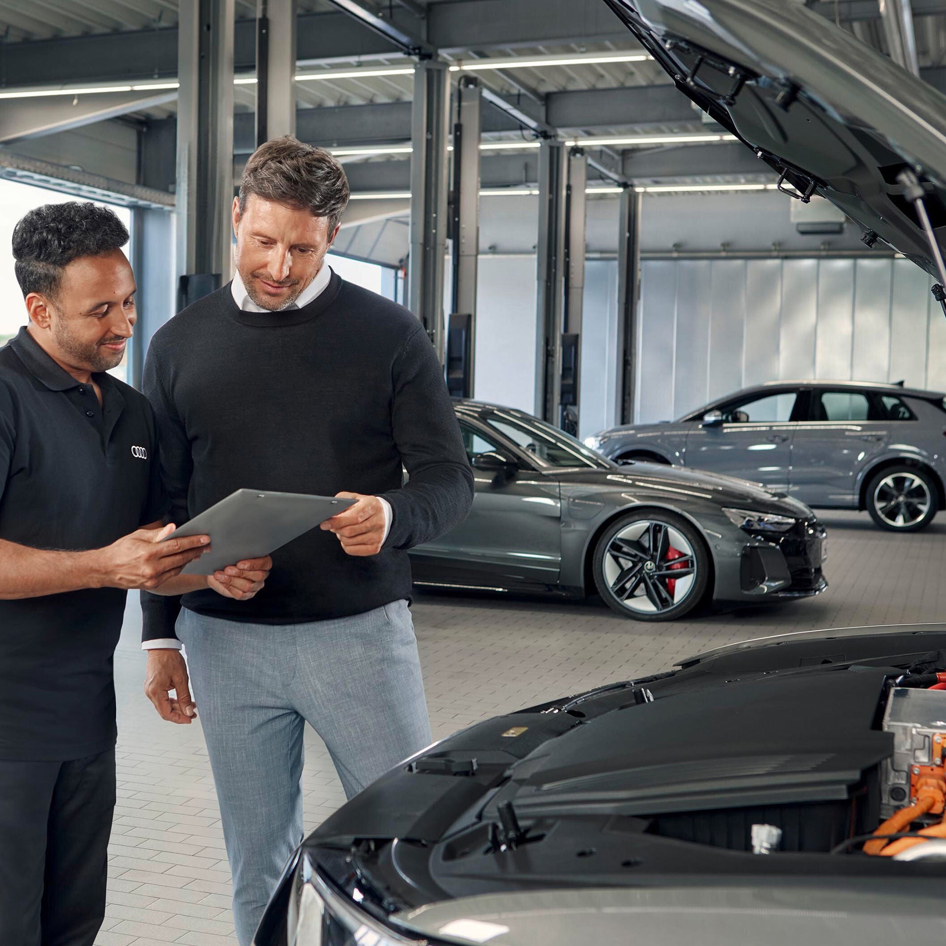 Servicemitarbeiter zeigt Kunden ein Tablet vor geöffnetem Auto in moderner Werkstatt mit zwei geparkten Audi-Fahrzeugen.