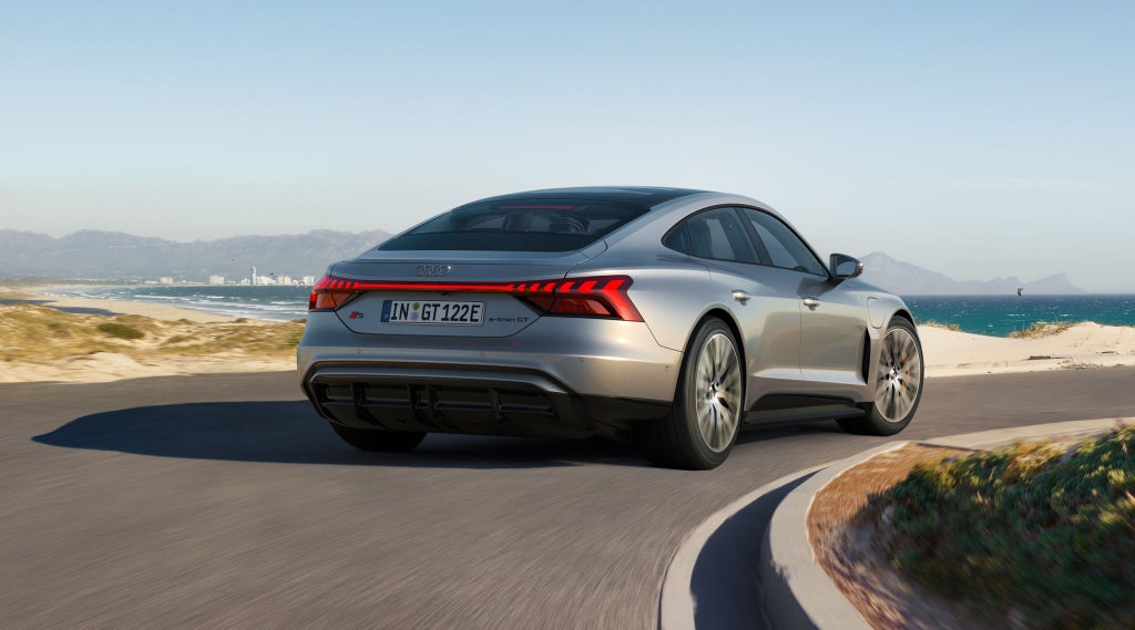 Ein silberner Audi S e-tron GT fährt auf einer kurvigen Küstenstraße mit Blick auf das Meer und eine Stadt im Hintergrund.