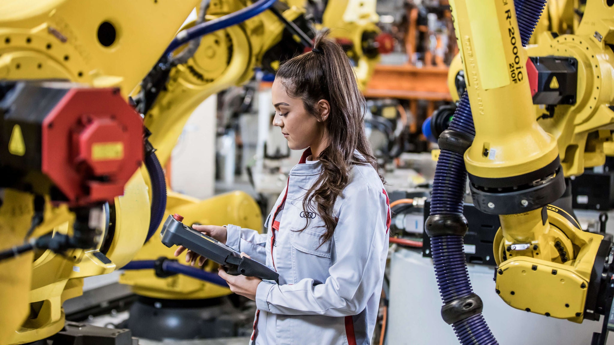 Frau in Audi-Uniform steuert Roboter in industrieller Fertigungshalle.