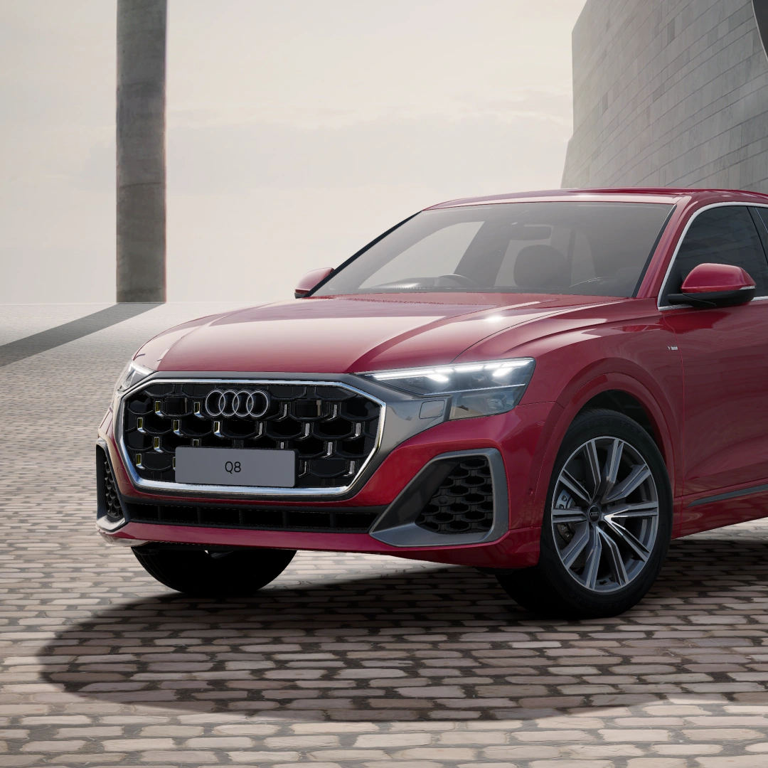 Front three-quarter view of a red Audi Q8 e-hybrid SUV in S line trim, parked on cobbled paving, with a chrome-framed Singleframe grille.