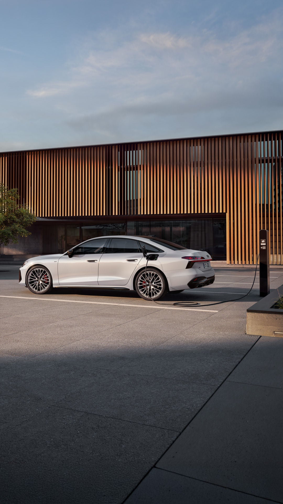Seitenprofil der weißen A6 Limousine e-hybrid, welche während des Ladens vor einem modernen Gebäude steht.